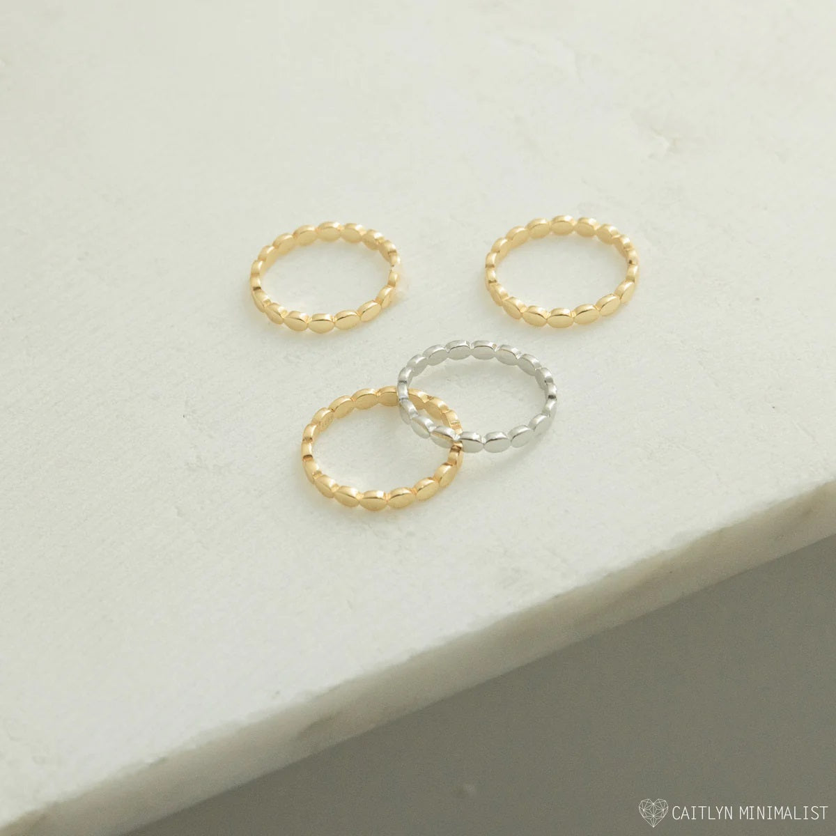Three gold and silver rings on a light gray surface with 'Caitlyn Minimalist' branding.