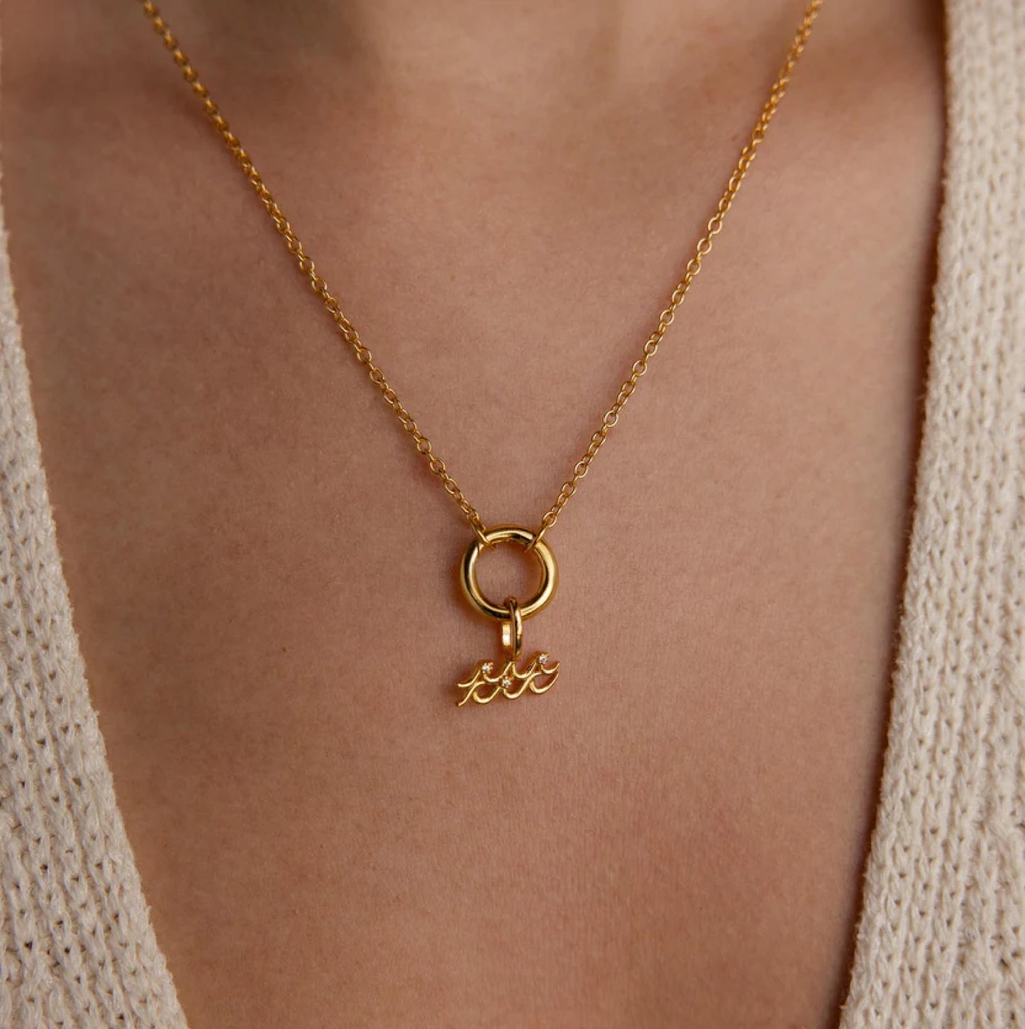 Gold necklace with a pendant on a person wearing a beige cardigan.