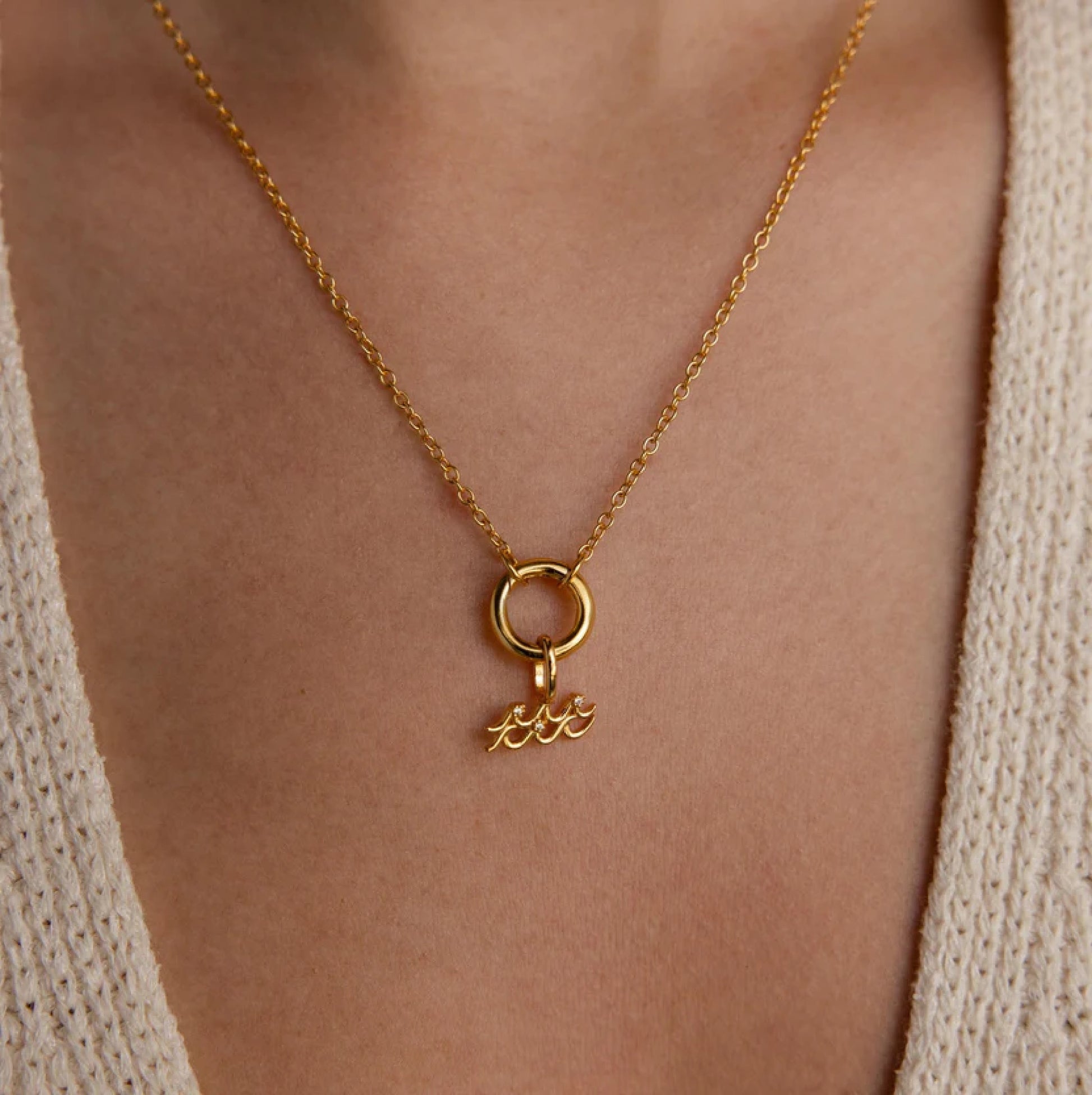 Gold necklace with a pendant on a person wearing a beige cardigan.