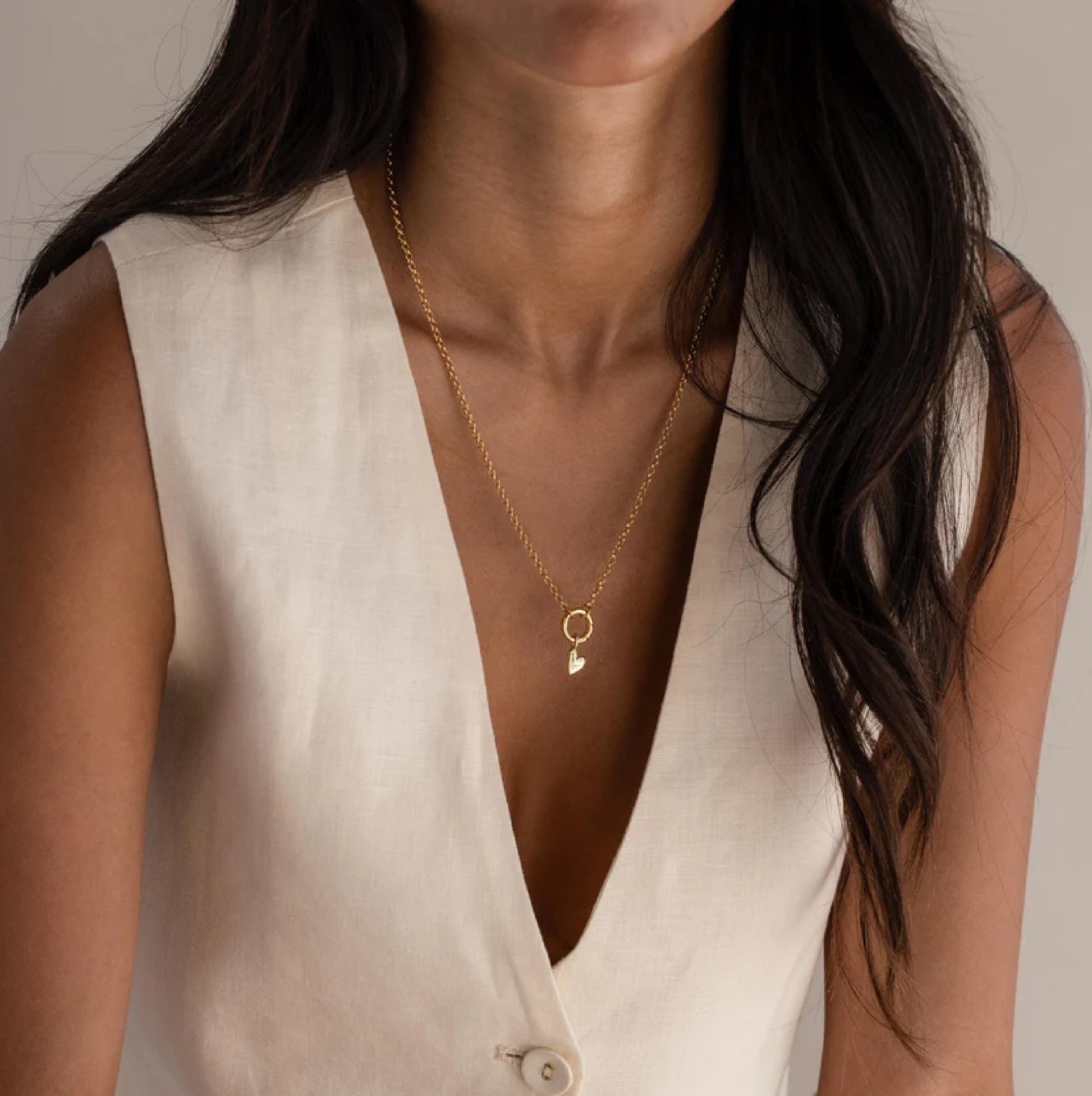 Close-up of a person wearing a gold necklace with a heart pendant on a neutral background