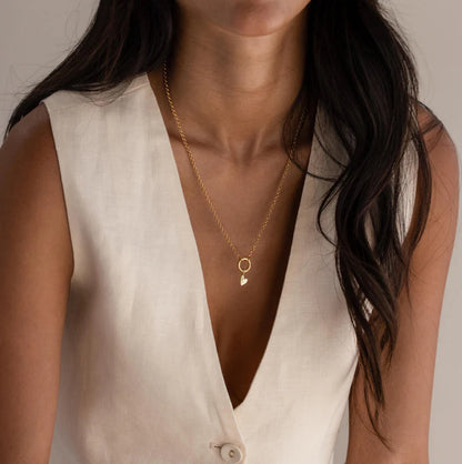 Close-up of a person wearing a gold necklace with a heart pendant on a neutral background