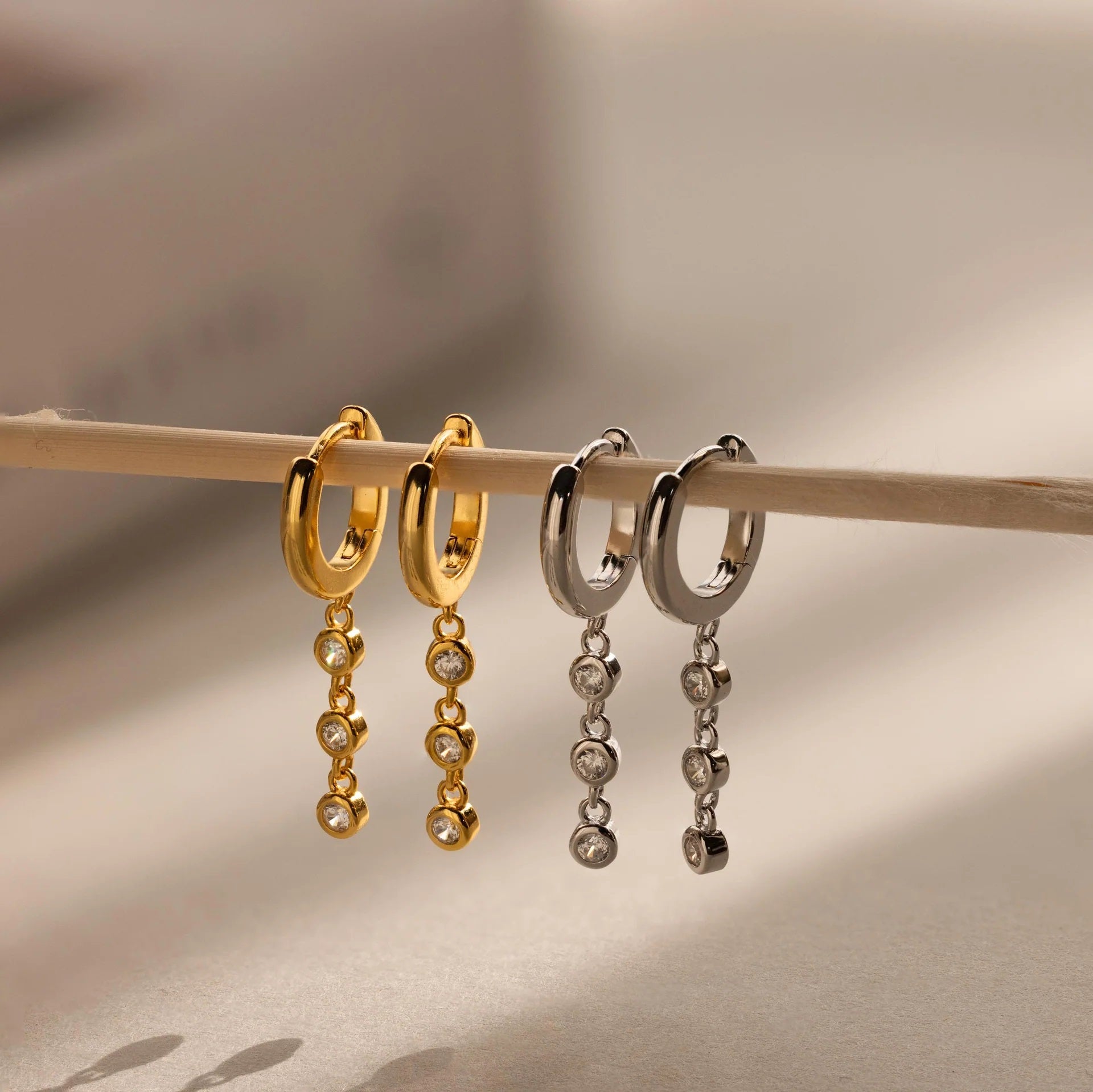 Gold and silver hoop earrings with dangling beads on a wooden stick against a neutral background.