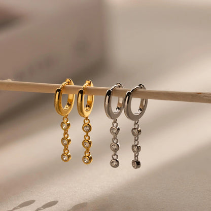 Gold and silver hoop earrings with dangling beads on a wooden stick against a neutral background.