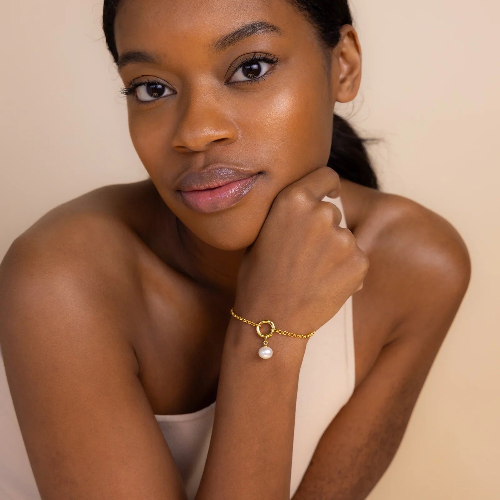 Woman wearing a gold bracelet with a pearl charm on a beige background