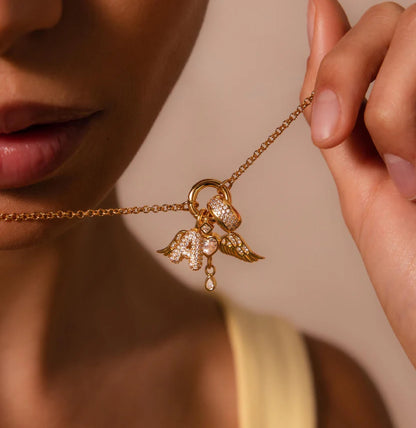 Gold necklace with angel wing and letter 'A' charm held by a hand against a neutral background