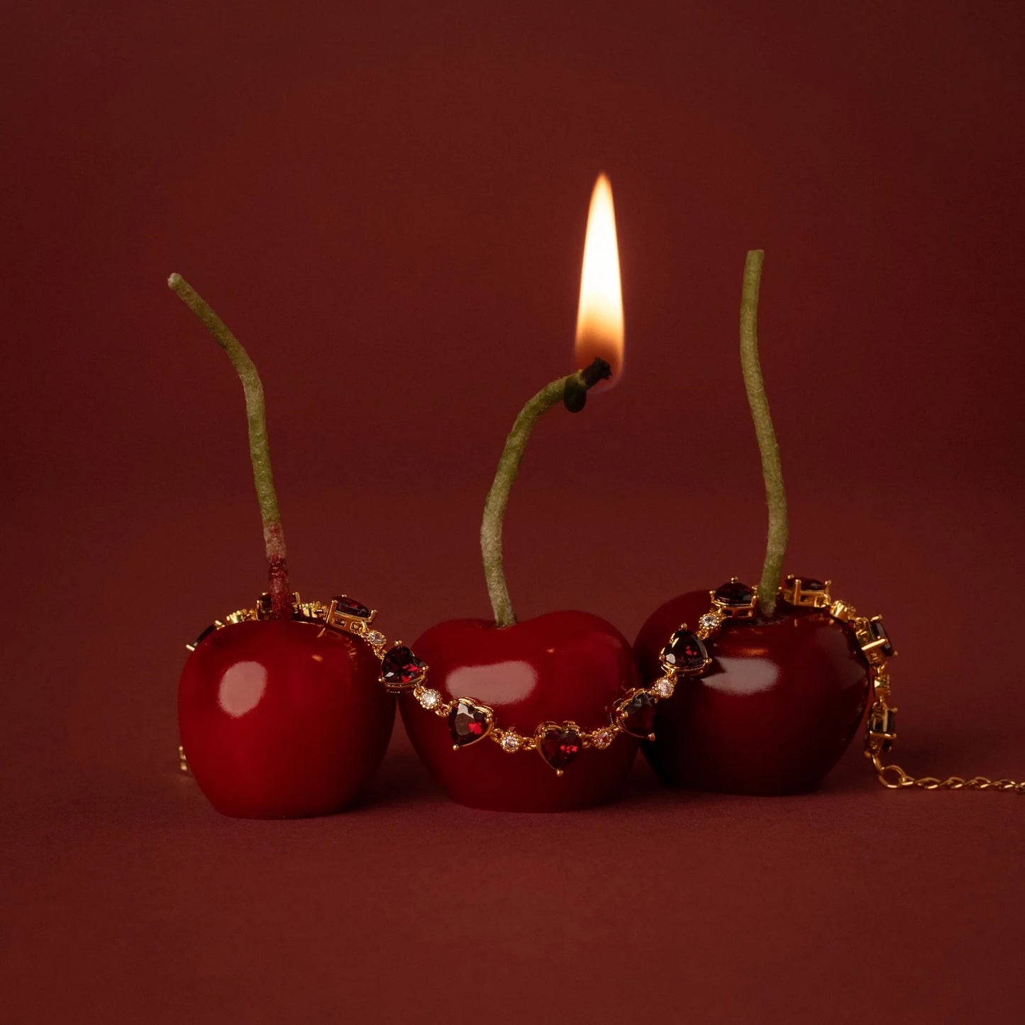 Three cherries with gold chains and a lit candle on a red background