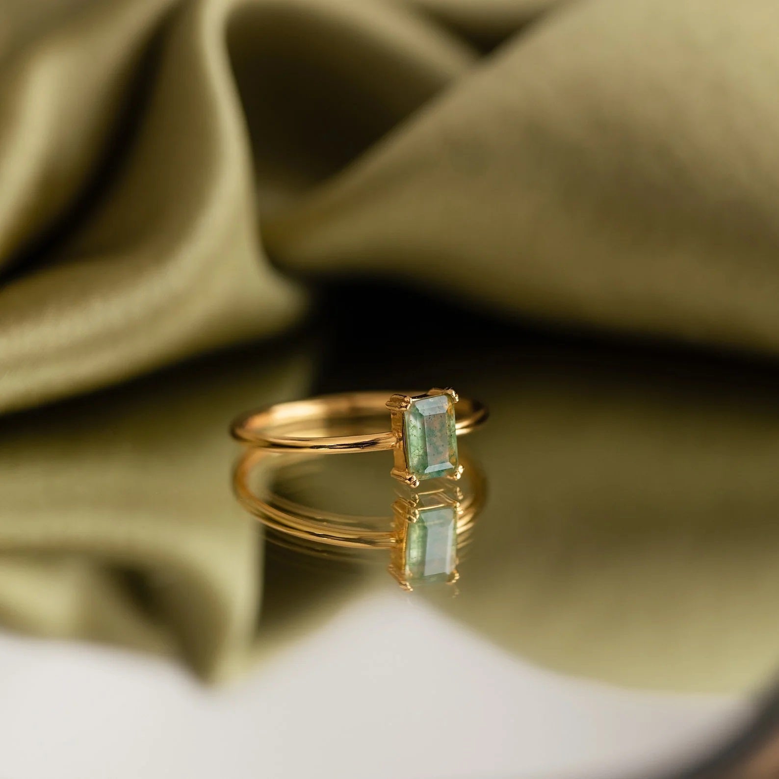 Gold ring with green gemstone on a reflective surface with green fabric background