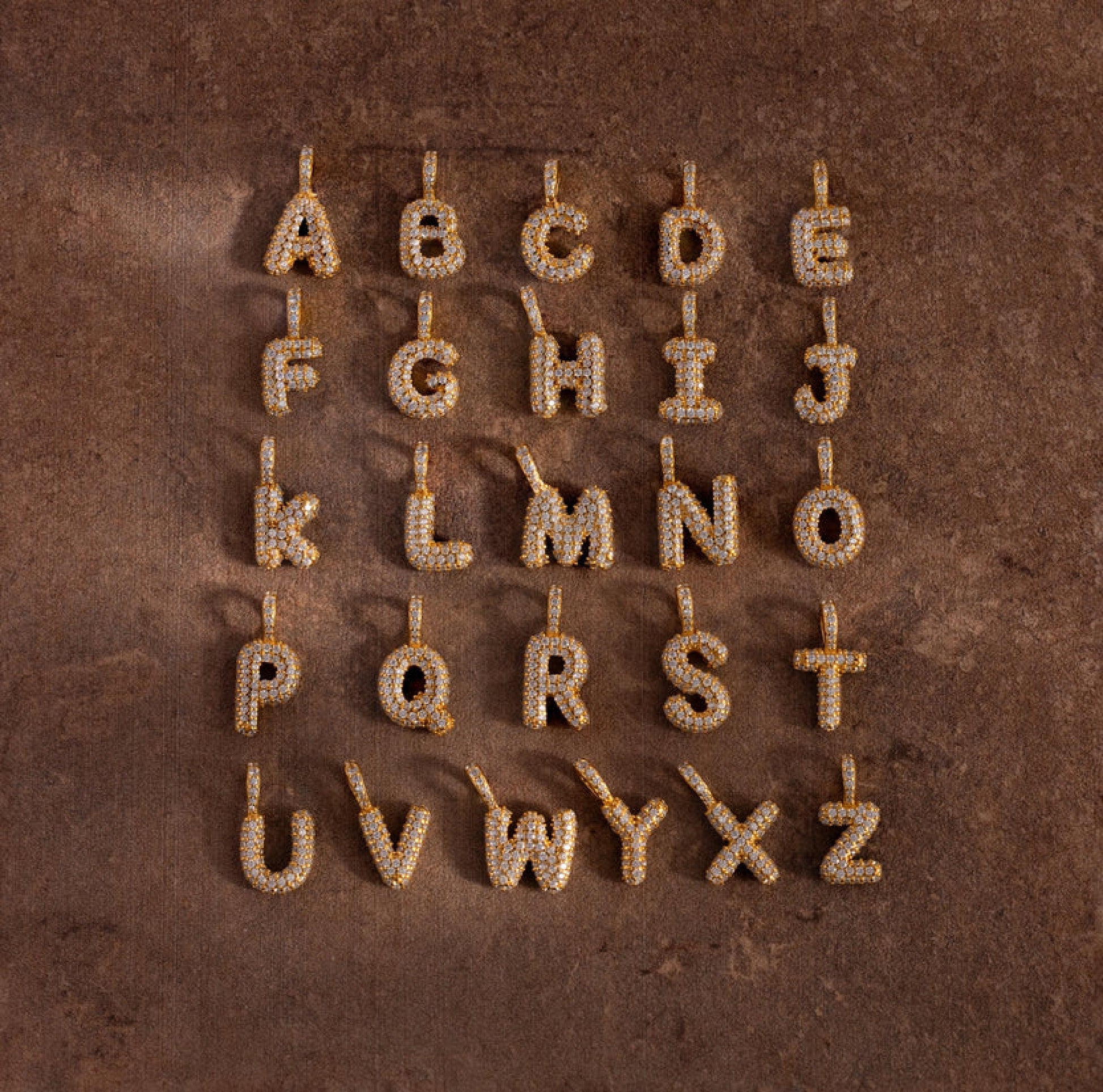 Gold letter earrings spelling out 'ABCDEFGHIJKLMNabcdefghijklm' on a brown background