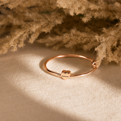 A delicate rose gold bangle bracelet with a small heart charm engraved with the word 'Gemma', resting on a textured beige fabric with dried pampas grass in the background.