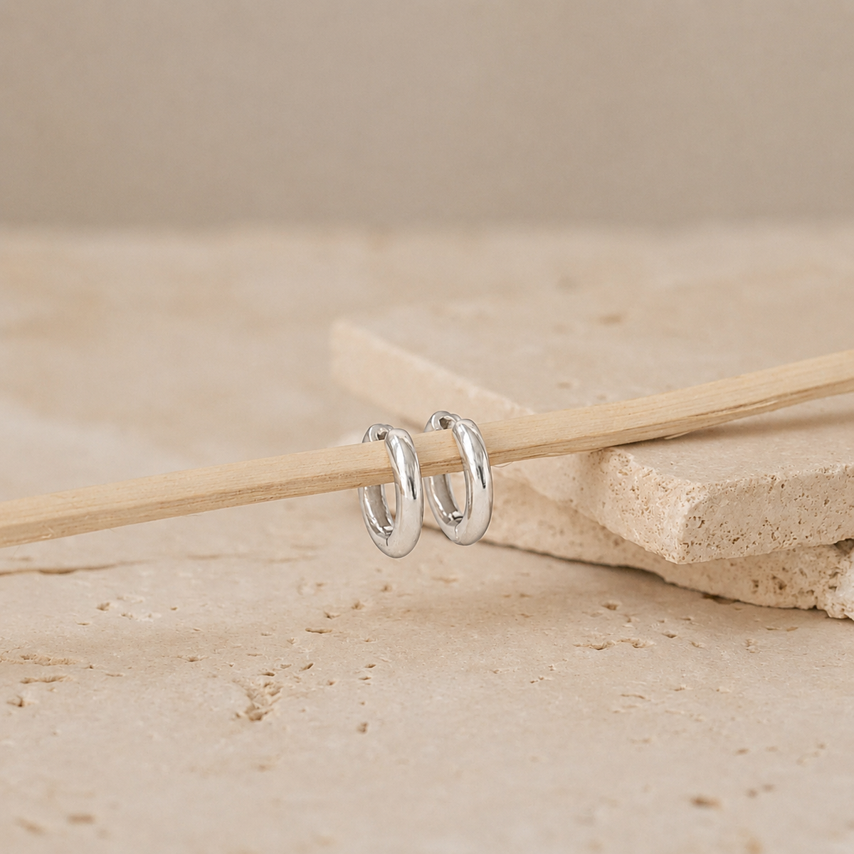 A pair of polished silver hoop earrings resting on a light wooden stick, set against a neutral beige stone background.