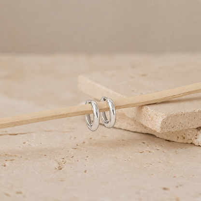 A pair of polished silver hoop earrings resting on a light wooden stick, set against a neutral beige stone background.