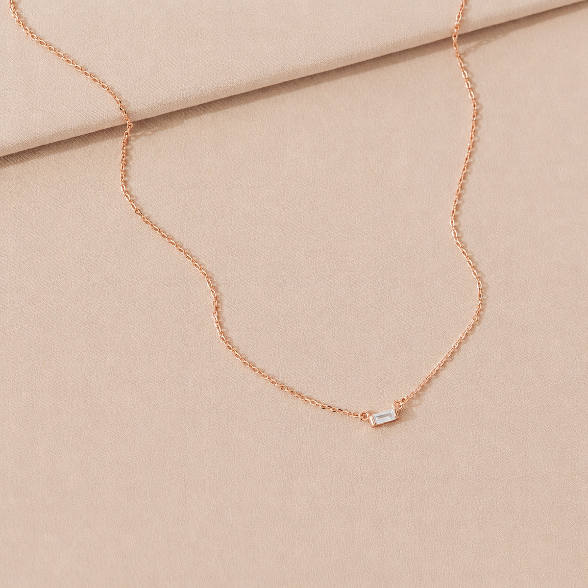 Rose gold necklace with a small rectangular pendant on a beige fabric background