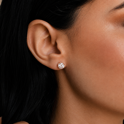 A close-up of a model's ear wearing a small round stud earring with a sparkling clear gemstone in a silver-toned setting.