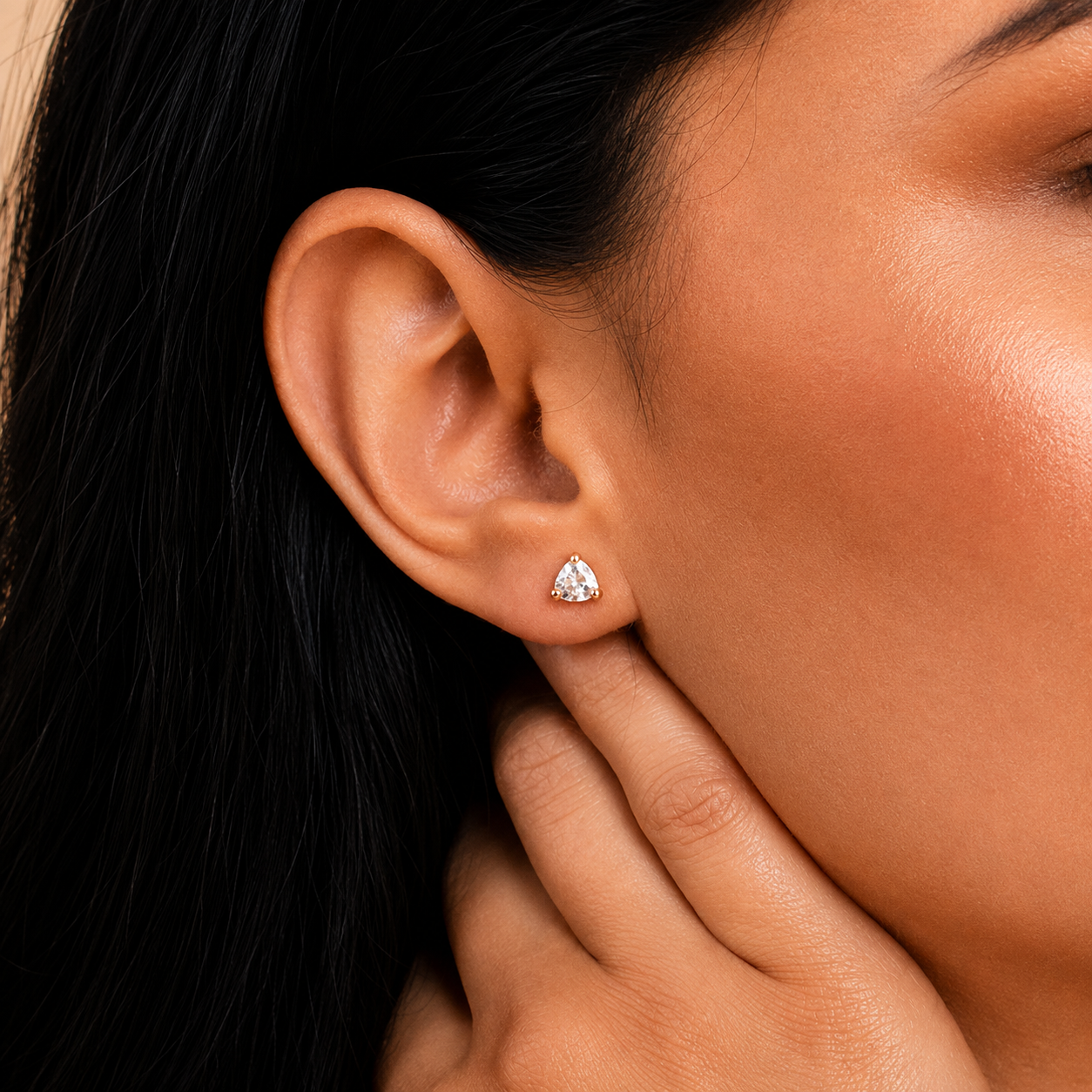 A close-up of a model's ear wearing a small triangular stud earring with a sparkling clear gemstone in a silver-toned setting.