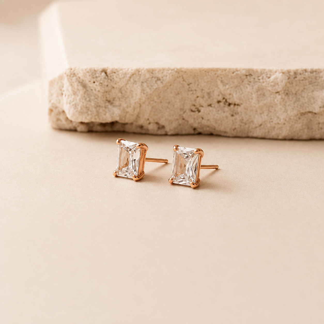 A pair of rose gold stud earrings with rectangular clear gemstones, resting on a beige surface with a textured stone prop in the background.