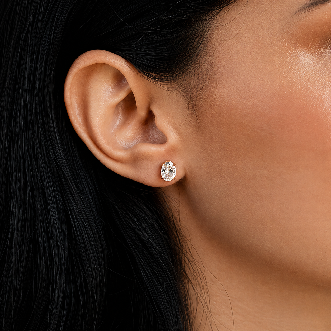 A close-up of a model's ear wearing a small oval stud earring with a clear gemstone in a silver-toned setting.