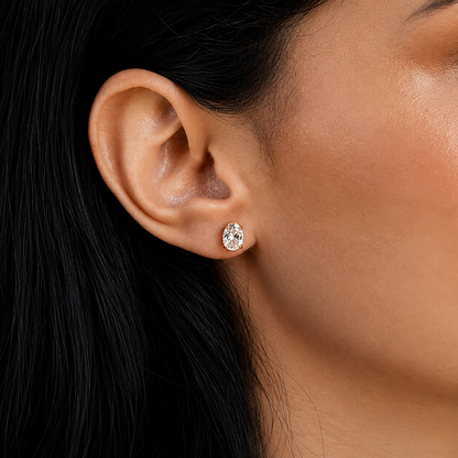 A close-up of a model's ear wearing a small oval stud earring with a clear gemstone in a silver-toned setting.