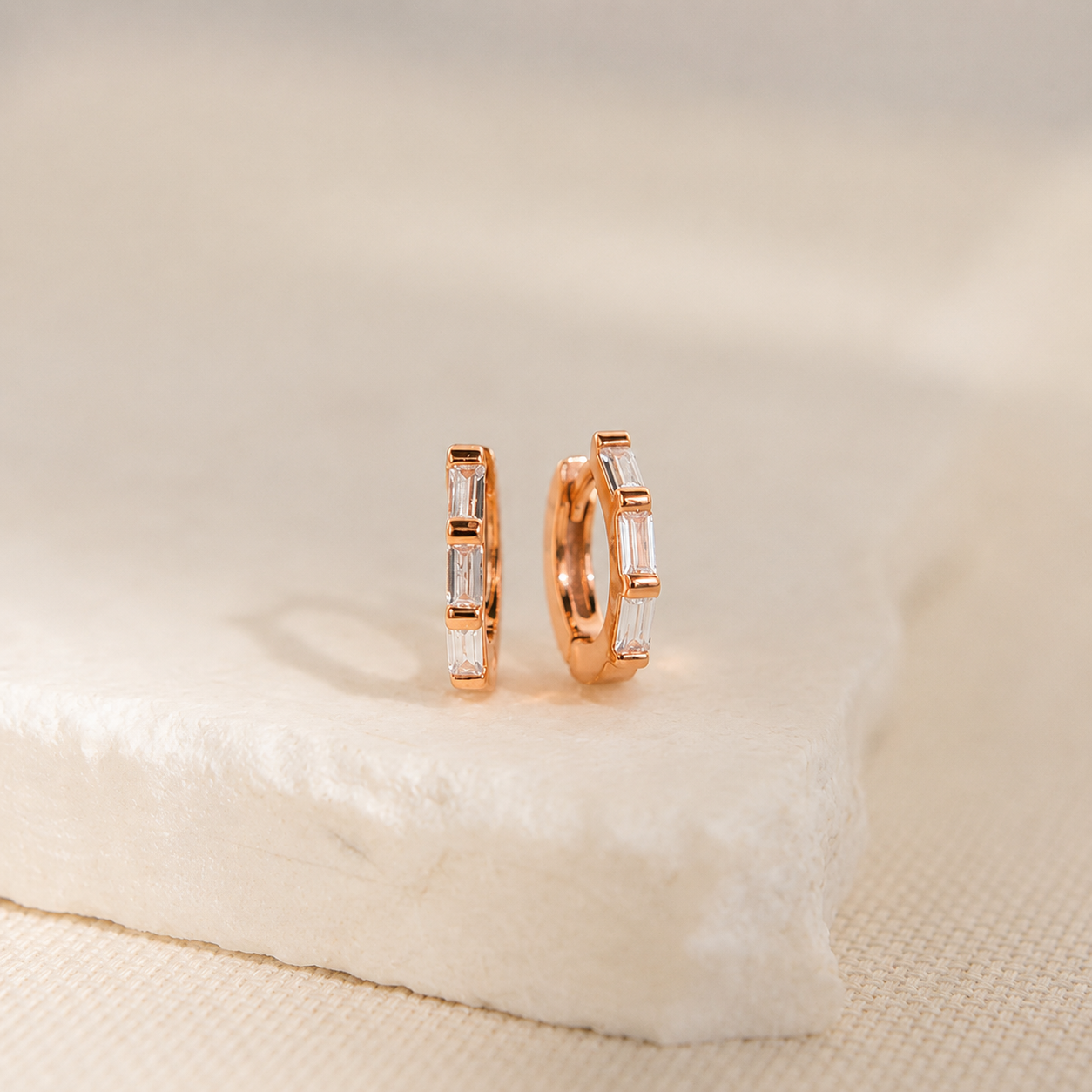 A pair of rose gold hoop earrings with baguette-cut clear stones, displayed on a textured white stone slab against a soft beige background.