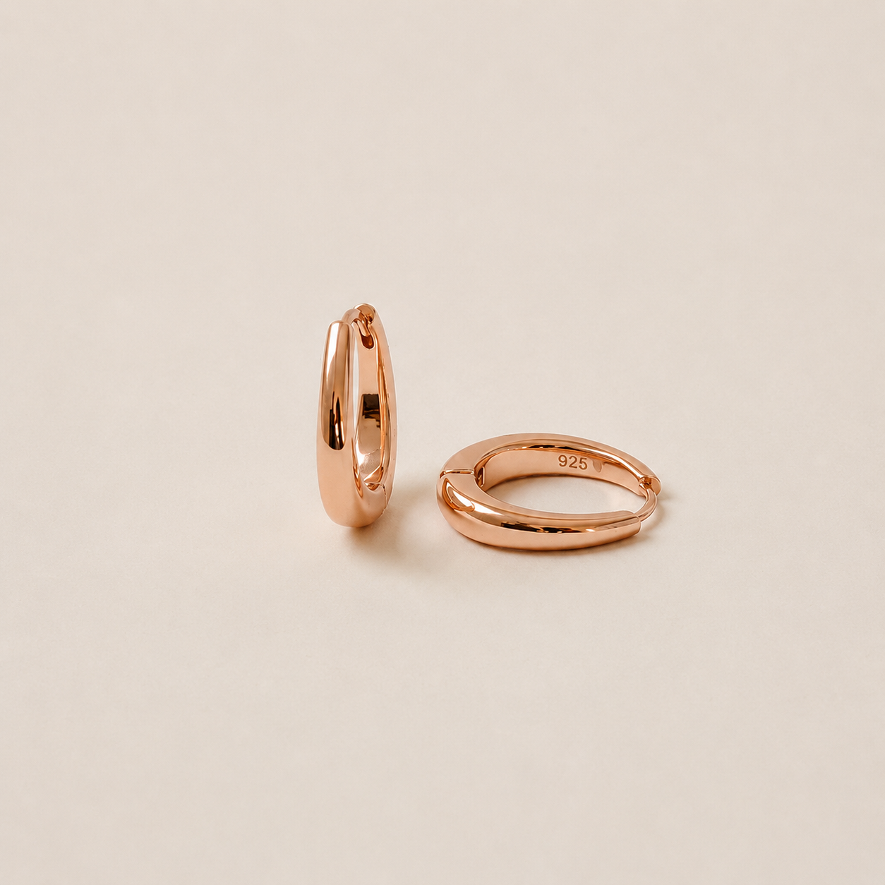 Two slim rose gold hoop earrings shown on a warm beige background, one standing vertically and the other lying horizontally. The horizontal hoop has a small '925' sterling silver purity stamp engraved on the inside.