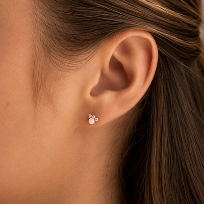 A close-up of a person's ear wearing a small stud earring with a pink gemstone and a tiny bow-shaped accent in a gold-tone setting.