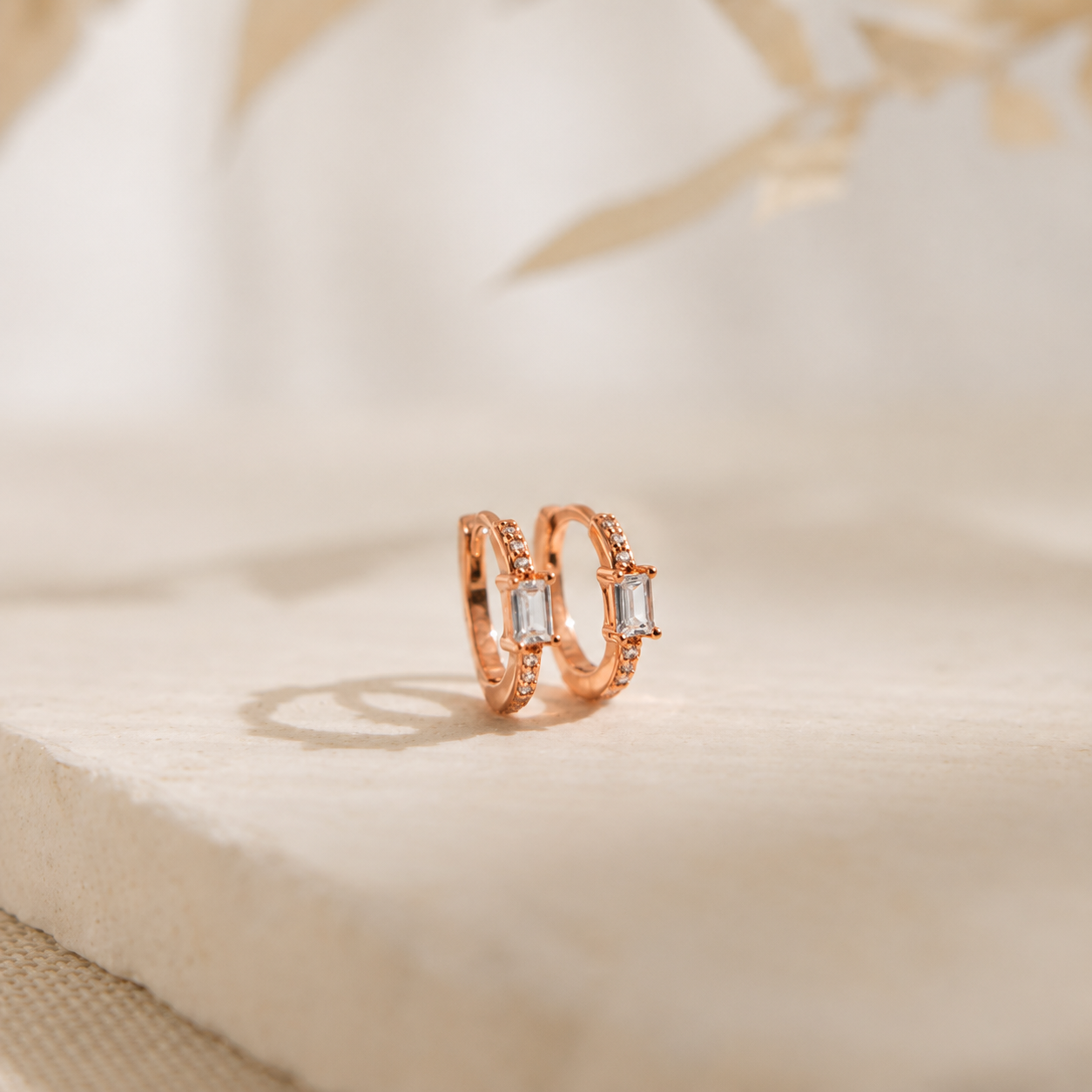 A pair of rose gold rings with rectangular clear gemstones, resting on a textured beige stone slab with a soft-focus background.
