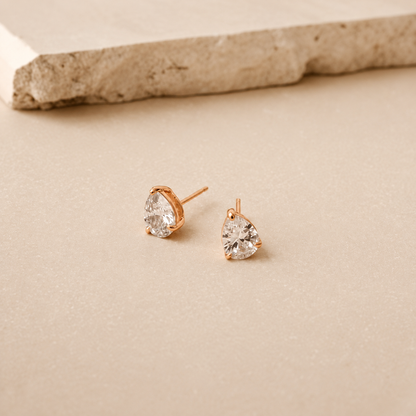 A pair of rose gold stud earrings with heart-shaped clear gemstones, resting on a textured beige surface with a stone prop in the background.