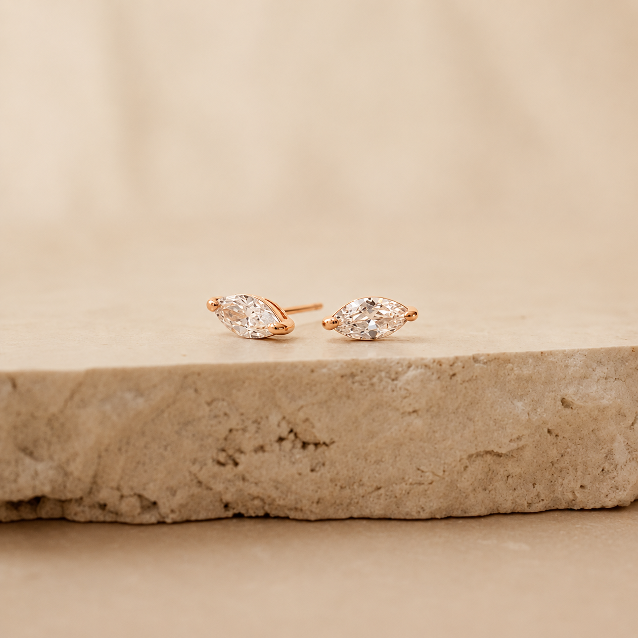 A pair of rose gold stud earrings with marquise-cut clear gemstones, resting on a textured beige stone slab against a soft beige background.