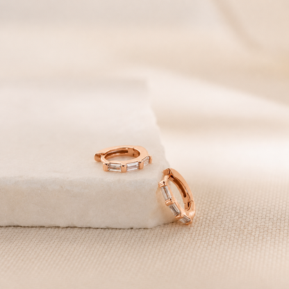 A pair of rose gold huggie hoop earrings with clear baguette-cut crystals, displayed on a white textured velvet block over a beige linen surface.