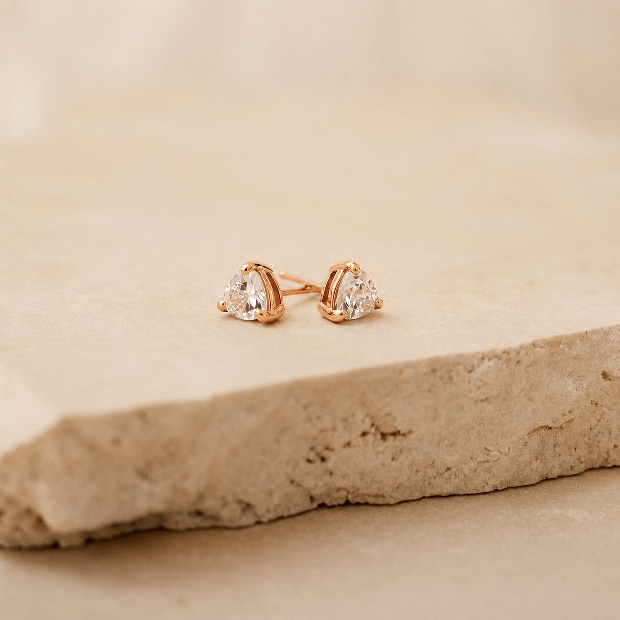 A pair of rose gold stud earrings with pear-cut clear gemstones, resting on a textured beige stone slab against a soft beige background.