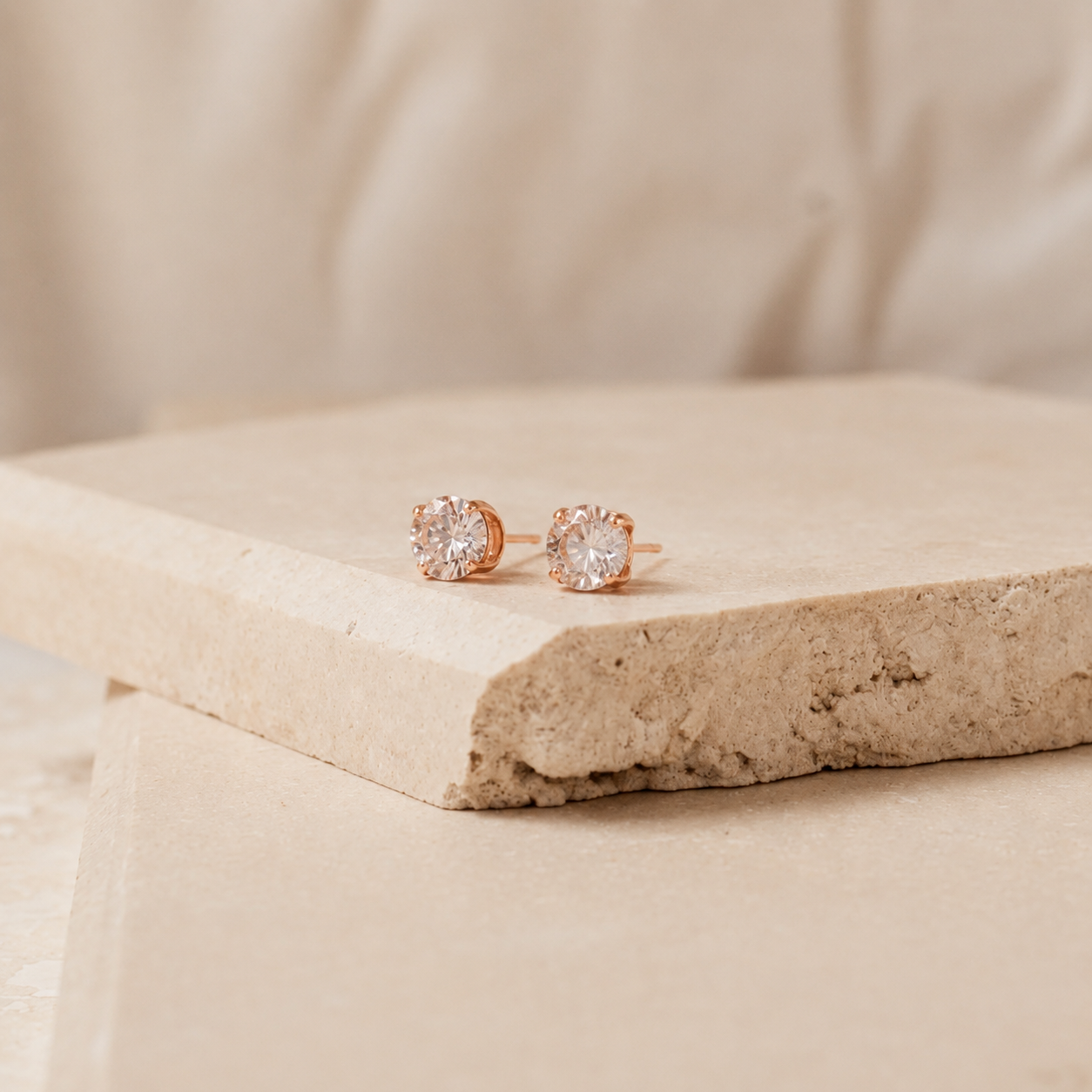 A pair of rose gold stud earrings with clear gemstones, resting on a textured beige stone slab with a soft beige fabric background.