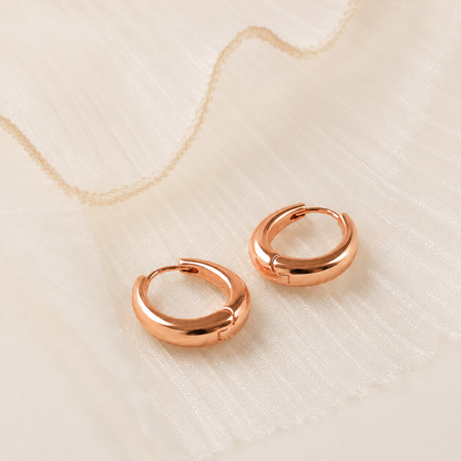 A pair of polished rose gold huggie hoop earrings resting on a textured, off-white fabric surface with a delicate gold chain in the background.