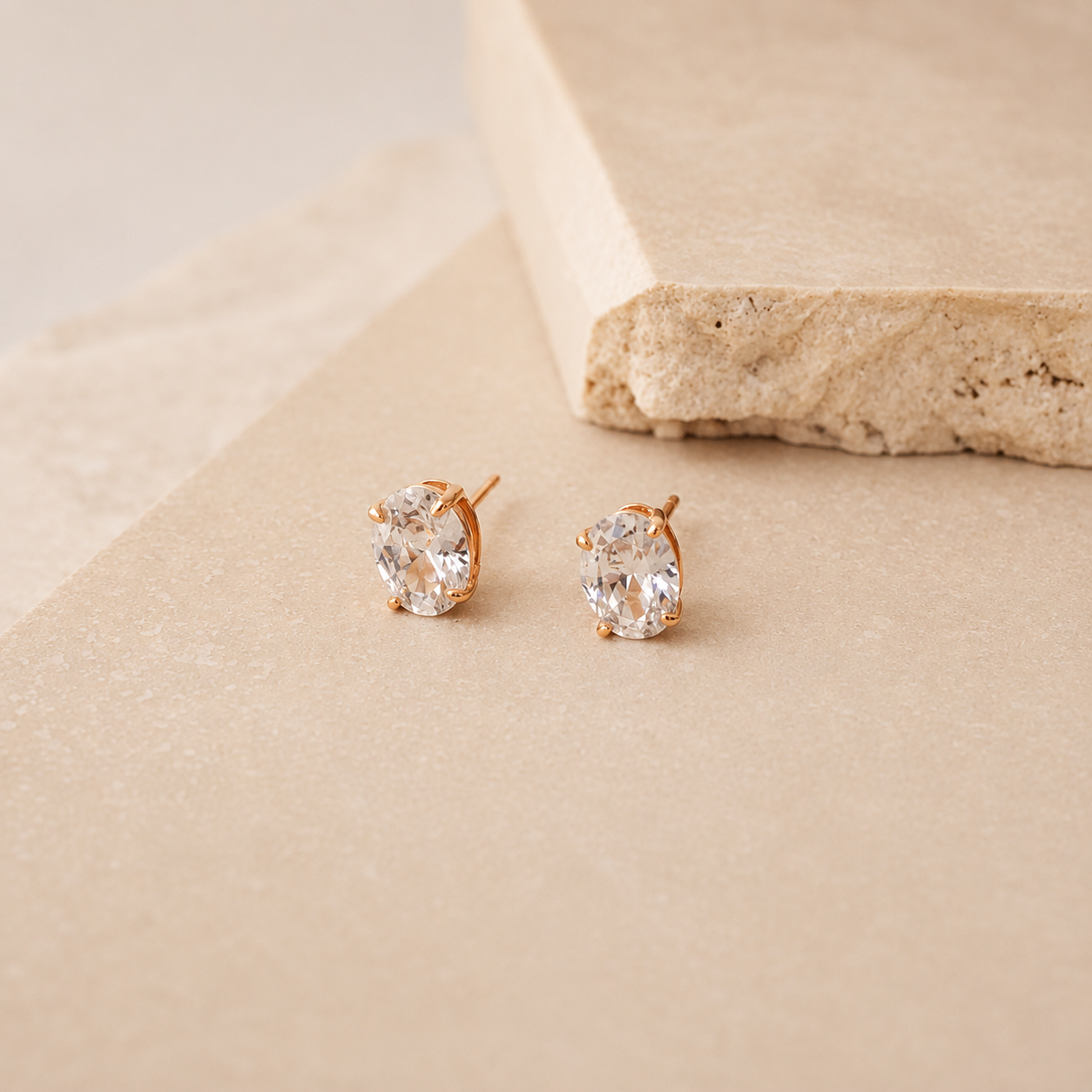 A pair of oval stud earrings with clear gemstones in a gold-tone setting, displayed on a textured beige surface next to a stone prop.