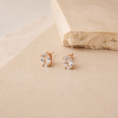 A pair of oval stud earrings with clear gemstones in a gold-tone setting, displayed on a textured beige surface next to a stone prop.