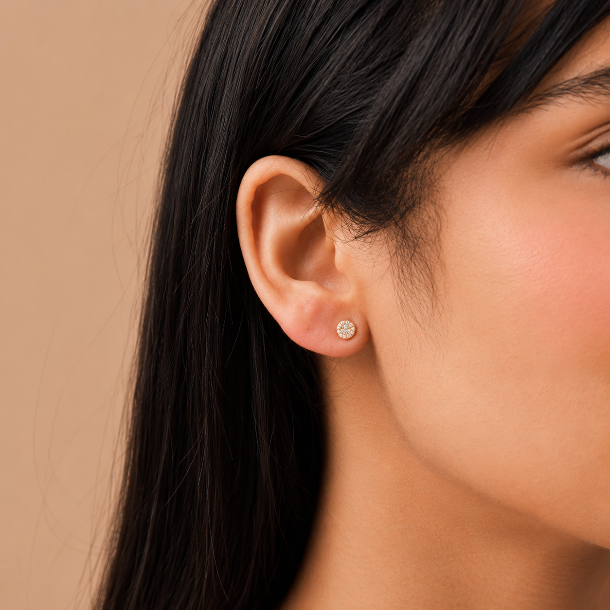 A close-up of a woman's ear wearing a small round stud earring with a silver-toned halo setting and clear stones, against a warm beige background.