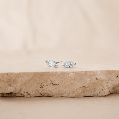 A pair of silver stud earrings with marquise-cut clear gemstones, resting on a textured beige stone slab against a soft beige background.