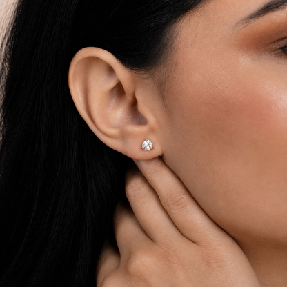 A close-up of a woman's ear wearing a small silver stud earring with a clear teardrop-shaped gemstone. The earring is shown on her earlobe, with her hand resting near her neck.