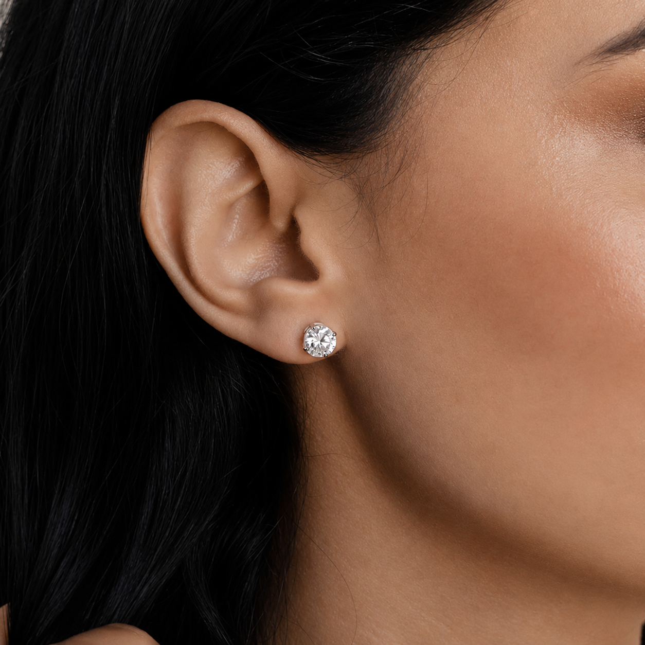 A close-up of a person's ear wearing a small round stud earring with a clear faceted gemstone in a silver-toned setting.