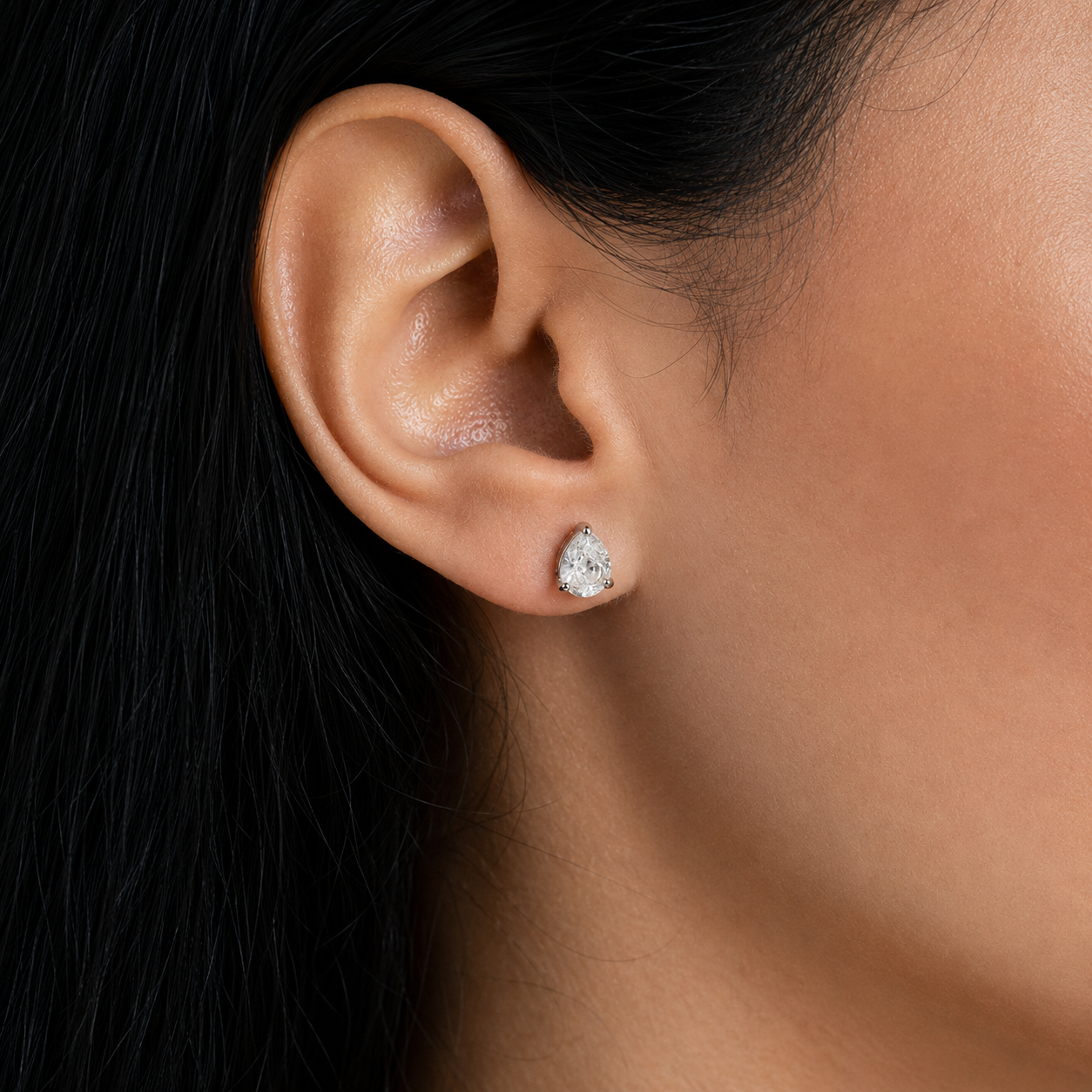 A close-up of a person's ear wearing a small teardrop-shaped clear gemstone stud earring in a silver setting.