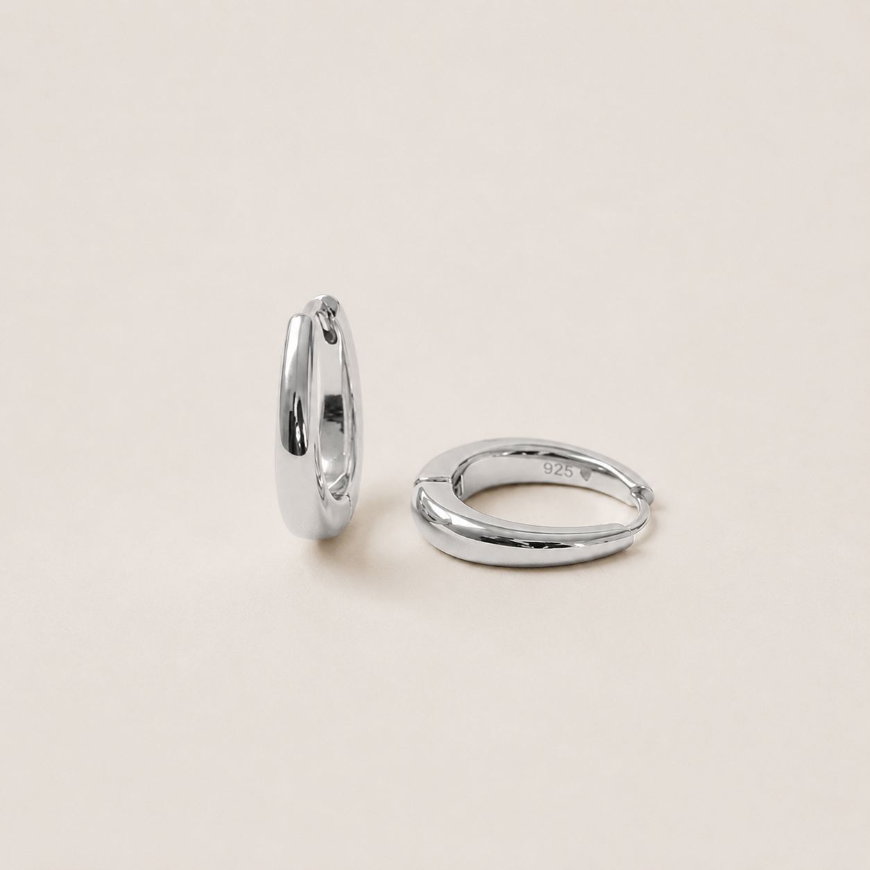 Two polished silver huggie hoop earrings, one standing vertically and the other lying horizontally, on a solid light beige background.