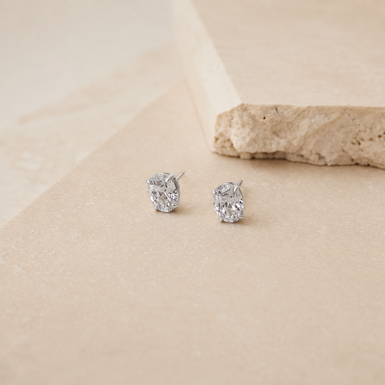 A pair of silver stud earrings with clear oval-cut gemstones, displayed on a textured beige surface with a stone prop in the background.