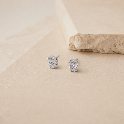 A pair of silver stud earrings with clear oval-cut gemstones, displayed on a textured beige surface with a stone prop in the background.