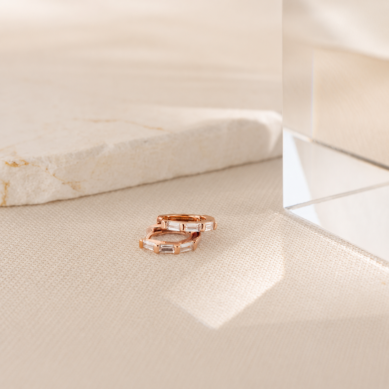 Two rose gold rings with baguette-cut clear stones, resting on a textured beige fabric surface next to a polished stone slab.