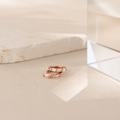 Two rose gold rings with baguette-cut clear stones, resting on a textured beige fabric surface next to a polished stone slab.
