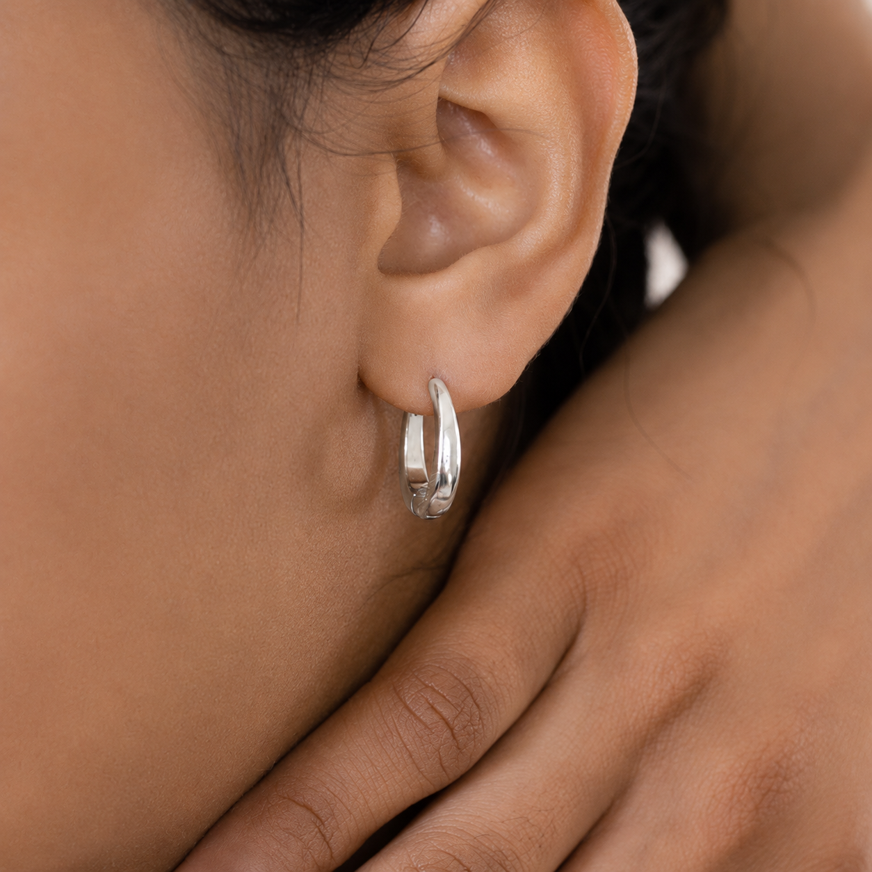 A close-up of a person's ear wearing a small polished silver hoop earring, with the hand resting near the jaw.
