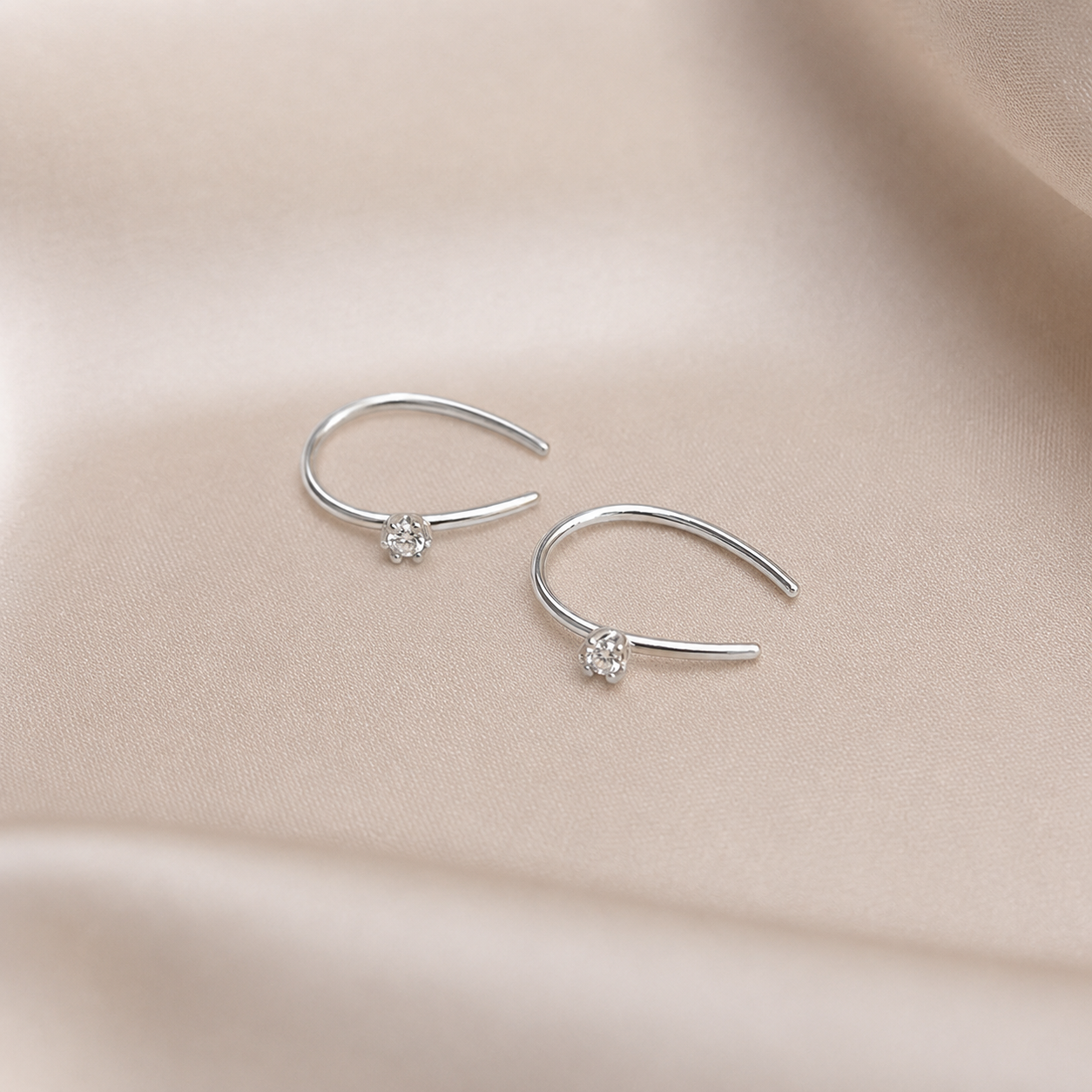 A pair of delicate silver ear cuffs with a small clear crystal flower setting, displayed on a soft beige satin fabric background.