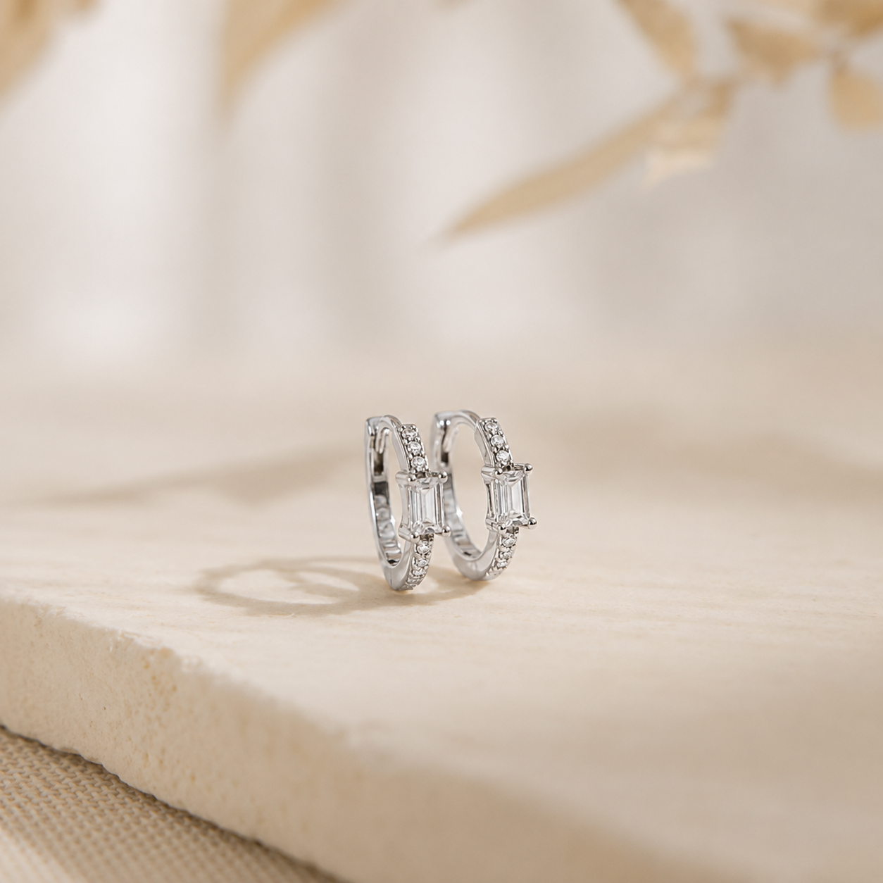 A pair of silver-toned rings with baguette-cut clear stones and small pavé accents, displayed on a light beige textured surface with a soft blurred background.