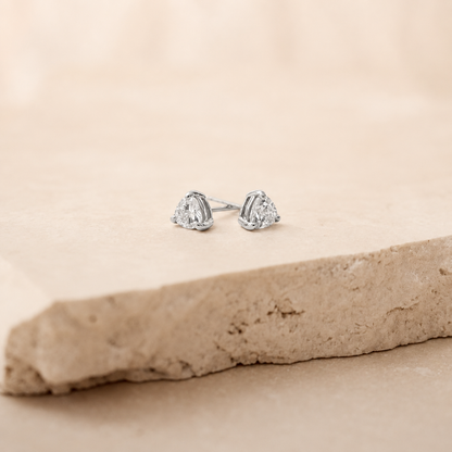 A pair of silver stud earrings with triangular clear gemstones, resting on a textured beige stone slab against a soft beige background.