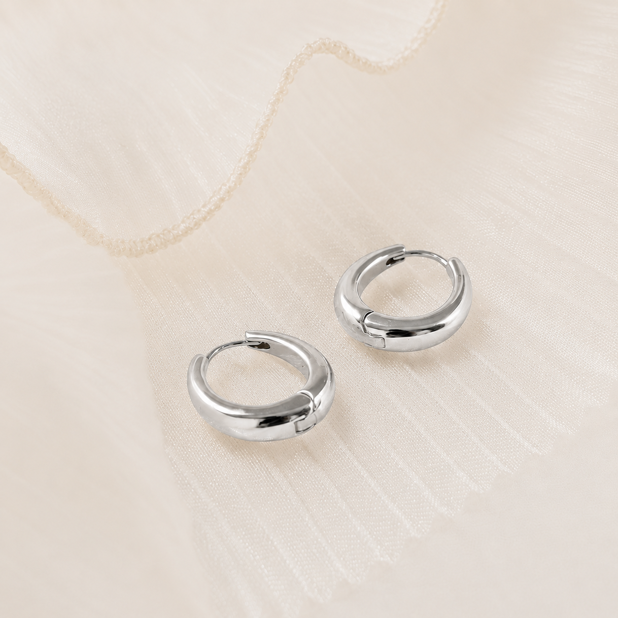 A pair of polished silver huggie hoop earrings resting on a textured, cream-colored fabric background.