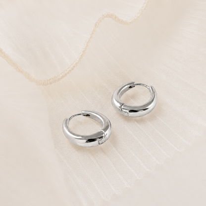A pair of polished silver huggie hoop earrings resting on a textured, cream-colored fabric background.