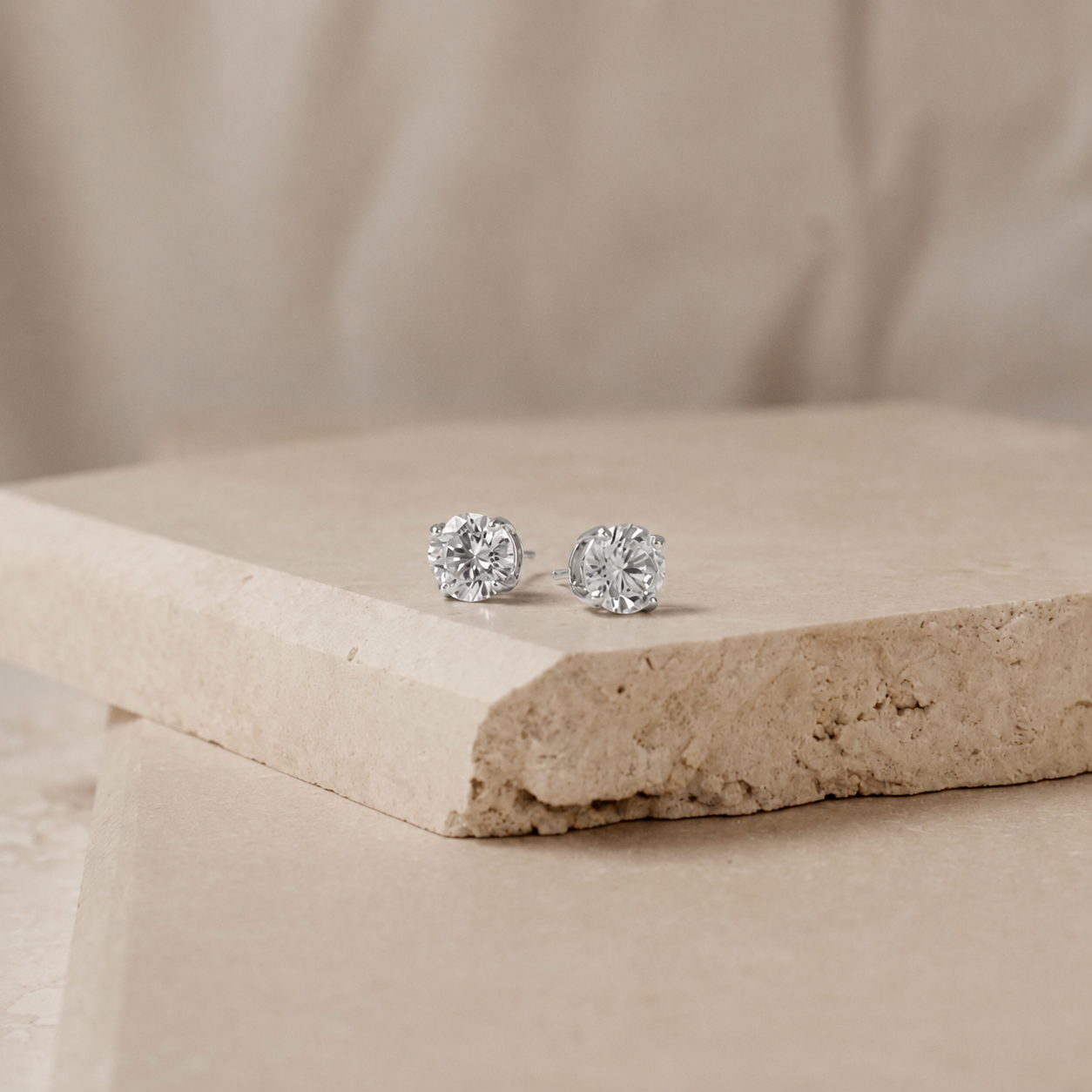 A pair of round-cut stud earrings with silver-toned settings, displayed on a textured beige stone slab against a soft beige fabric background.